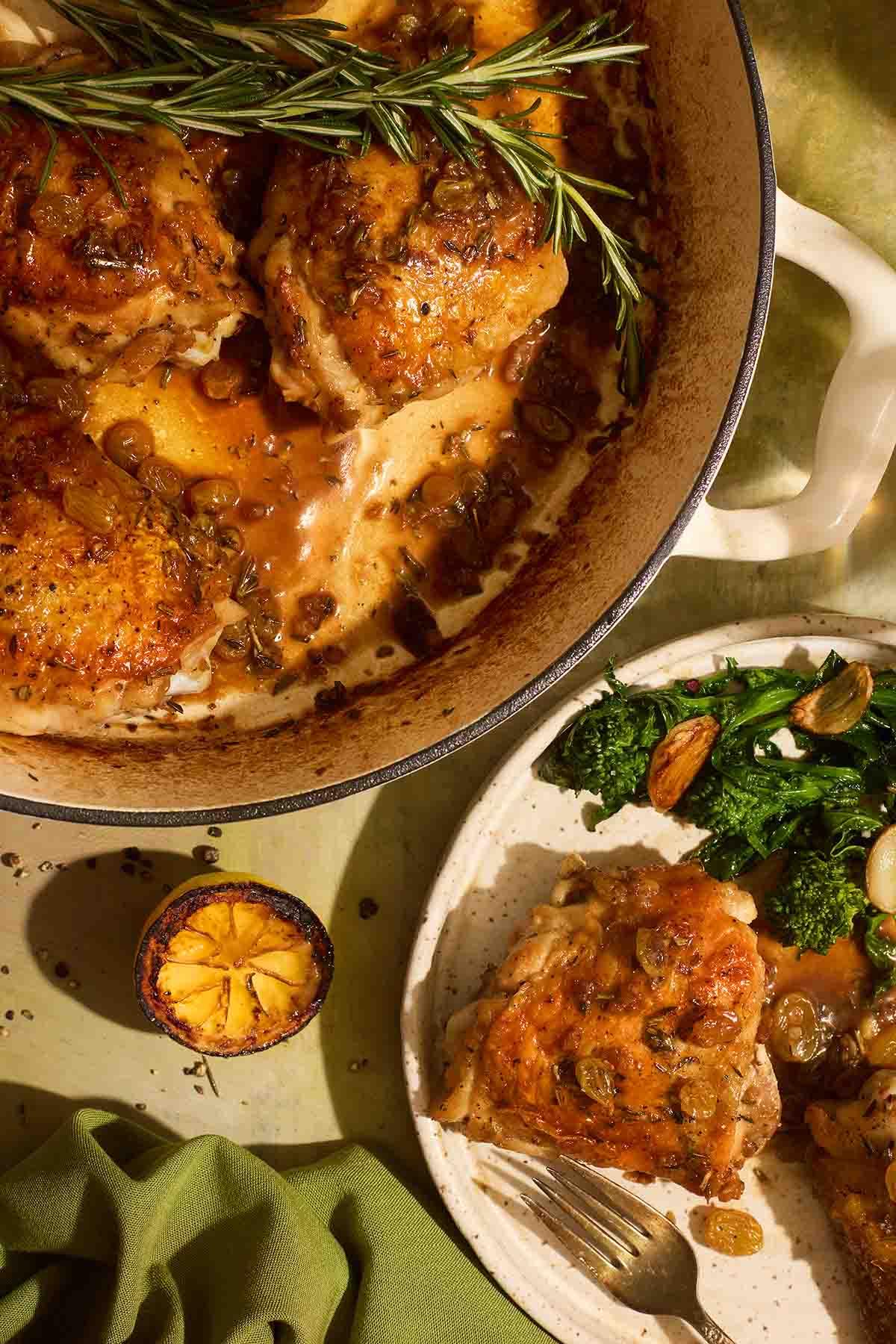 A white skillet of crispy chicken thighs covered in a glossy agrodolce sauce with golden raisins and a fresh rosemary sprig, placed next to a serving plate.