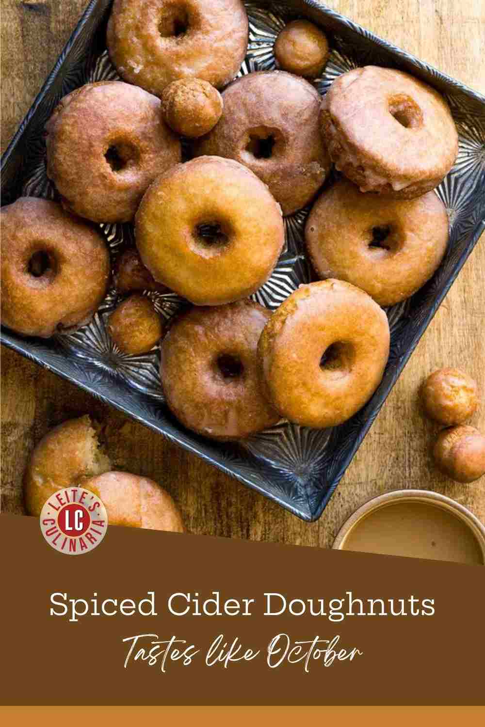 Glazed cider doughnuts and doughnut holes piled in a dark metal tray on wood, coffee alongside.