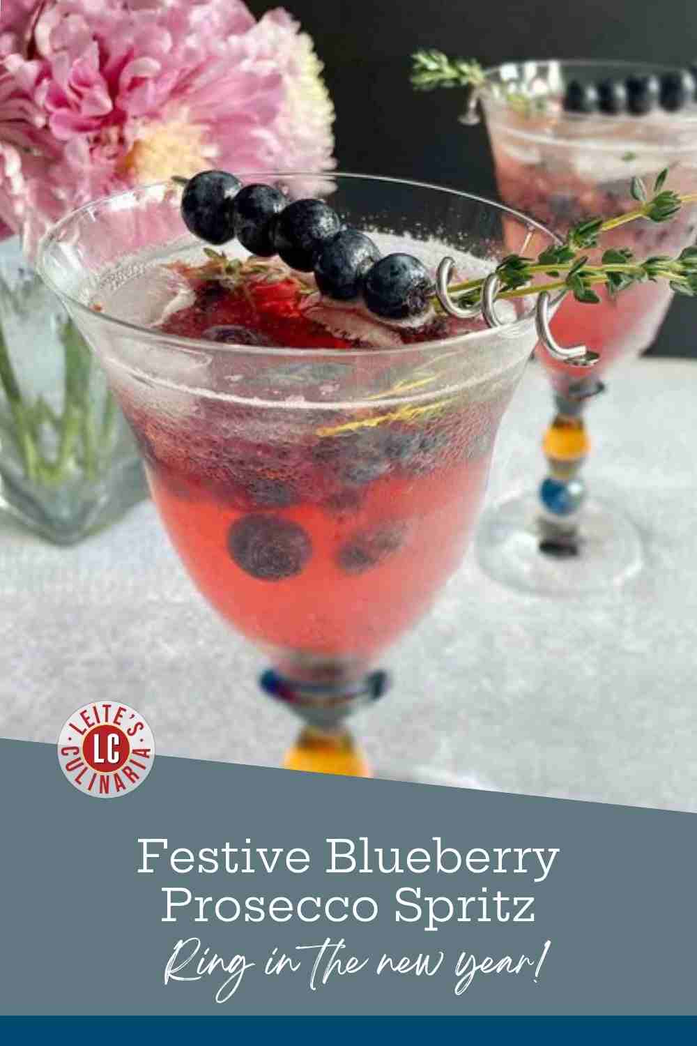 Two stemmed glasses of blueberry Prosecco spritz, one with blueberries and thyme skewered across the rim, beside pink dahlias.