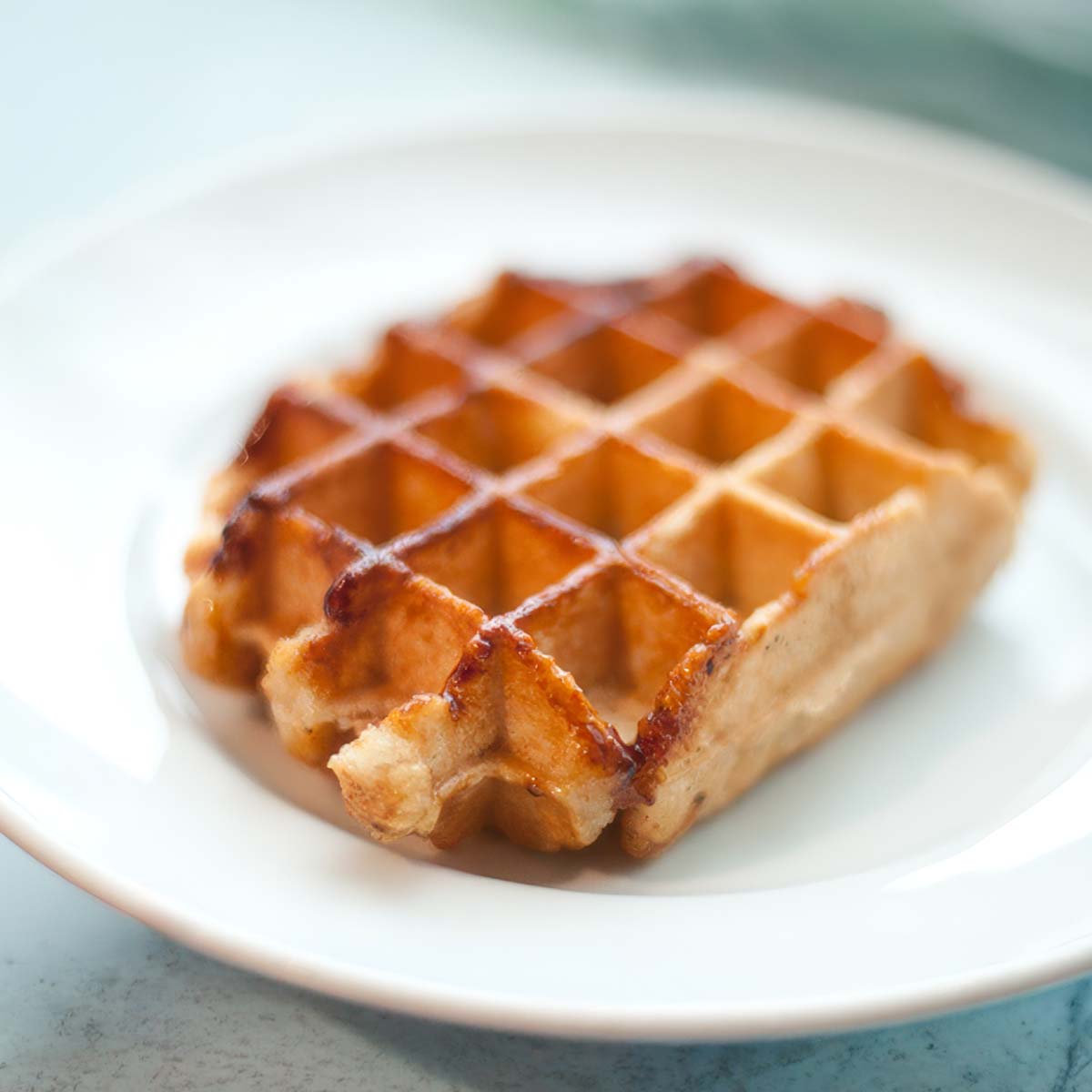 A single Liège waffle with a glossy, caramelized crust on a white plate over pale marble.