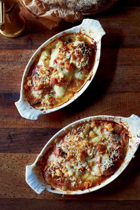 Golden-crusted endive gratin in two shell-handled dishes on dark wood, a brass pepper mill behind.