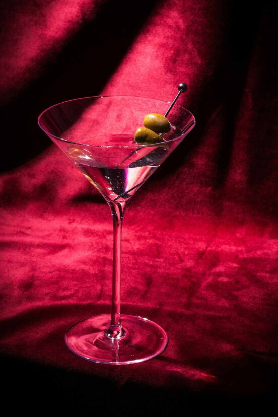 A long-stemmed martini glass filled to the rim, two green olives on a cocktail pick, against draped crimson velvet.