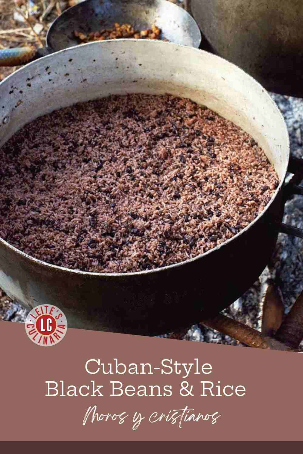 A large battered metal pot of Cuban black beans and rice cooking over a charcoal fire, with another pan behind it.