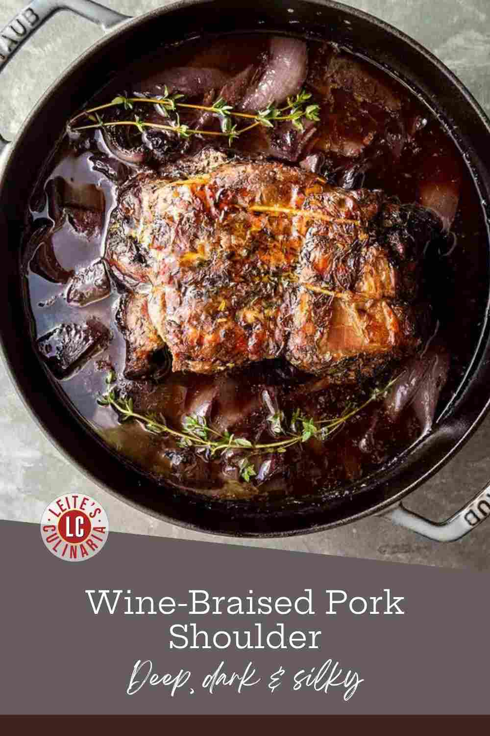 A whole braised pork shoulder in a Staub Dutch oven with red onions, dark wine sauce, and fresh thyme.