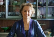 A Photo of Alice Waters standing in a kitchen smiling, wearing a blue sweater.