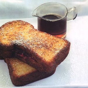 Two slices of cranberry French toast on a plate, sprinkled with icing sugar, in front on a pitcher of maple syrup.