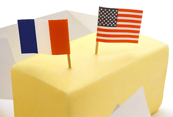 A stick of butter unwrapped with a small paper French flag and a small paper American flag both stuck in it.