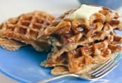 Gluten-free multigrain waffles piled on a plate, covered with melted butter and syrup.