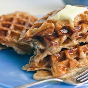 Gluten-free multigrain waffles piled on a plate, covered with melted butter and syrup.