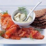 Gravadlax with sweet mustard piled on a plate with dill mayonnaise in a bowl, flanked by thinly sliced brown bread