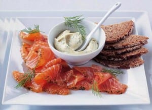 Gravadlax with sweet mustard piled on a plate with dill mayonnaise in a bowl, flanked by thinly sliced brown bread