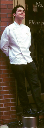 A chef in his chef's whites leaning up against a brick wall and smiling.